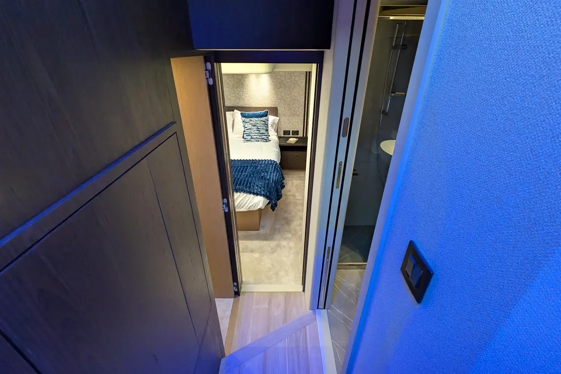 View from a hallway showing a stylish bedroom with a bed, blue throw, and ensuite bathroom, under soft lighting.